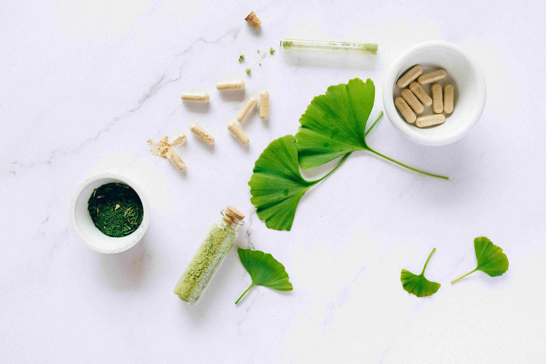 Green leaves, herbal powder and capsules in small white bowls on a pale grey surface